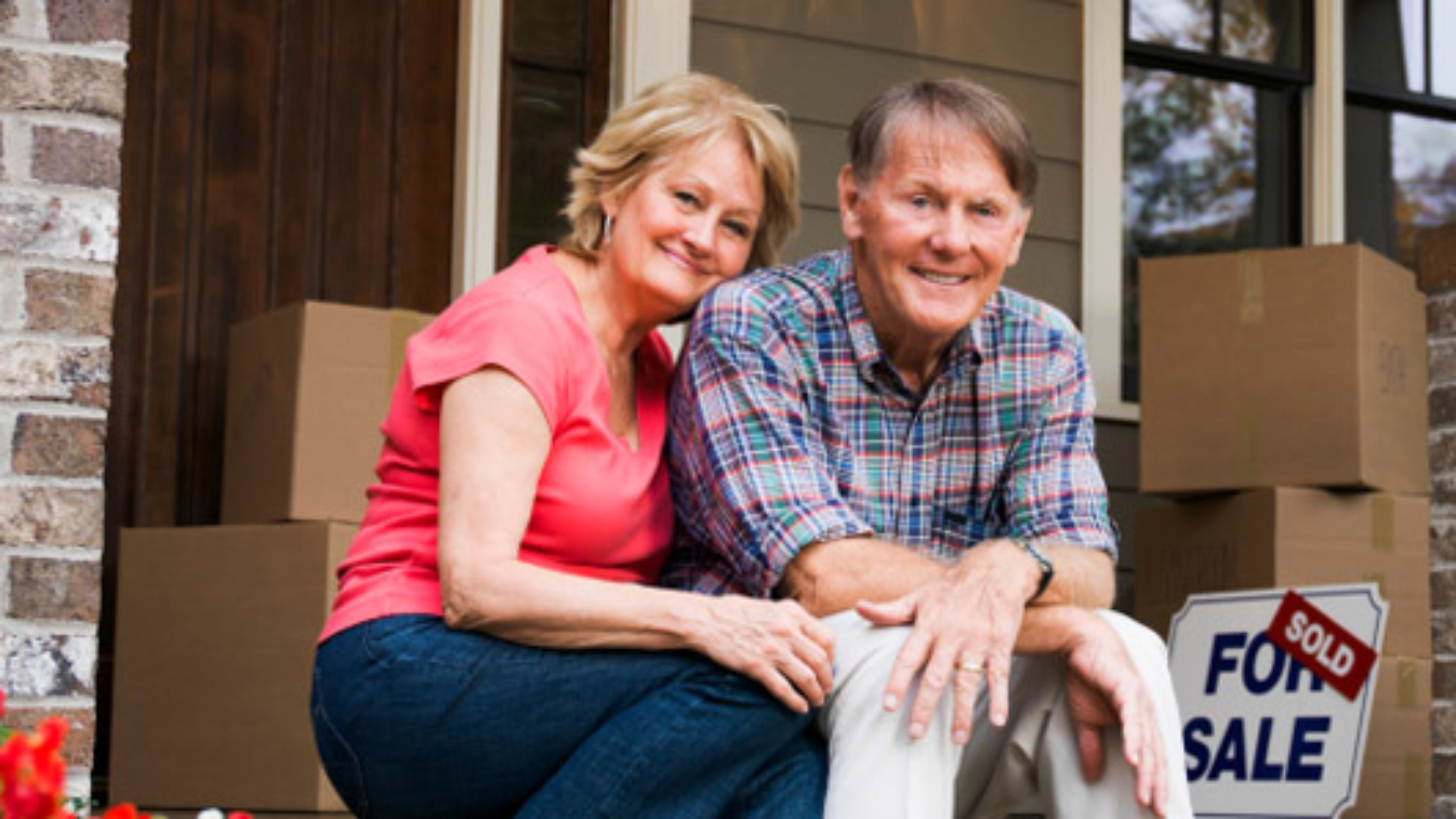 Portrait Of Happy Senior Couple Sitting Outside House With For Sale Sign